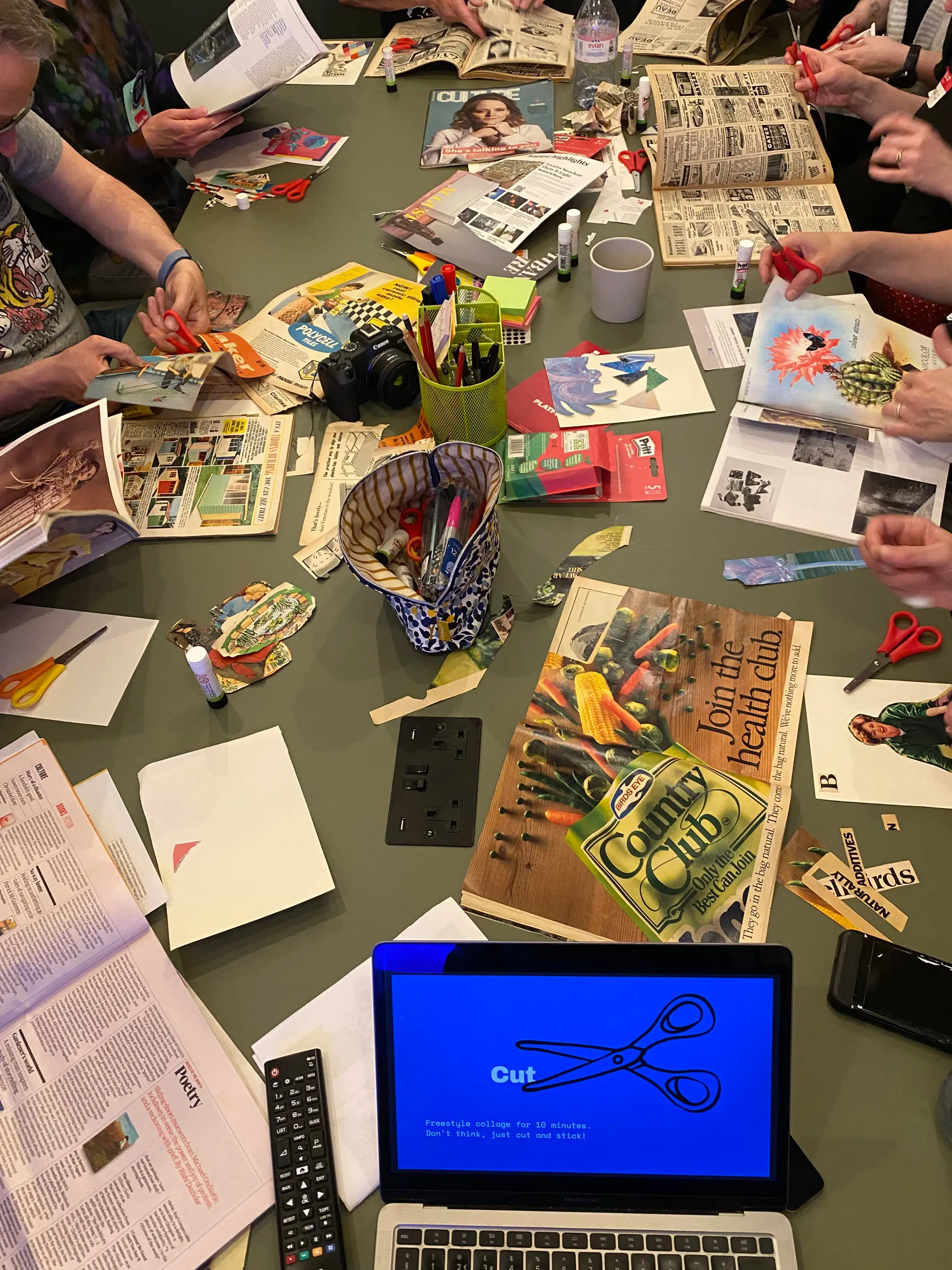 A table covered with paper, magazines, scissors, glue sticks and pens. In the foreground is a laptop. On the screen is an illustration of a pair of scissors and the word "cut".