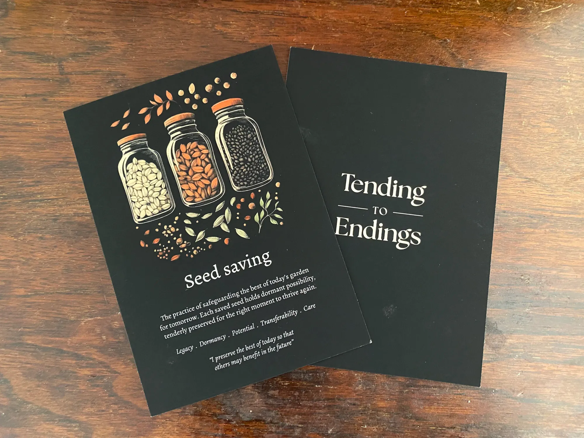 Two cards from the Tending to Endings card deck. The one on the left has an illustration of three jars of seeds. The text reads: "Seed saving. The practice of safeguarding the best of today's garden for tomorrow. Each saved seed holds dormant possibility, tenderly preserved for the right moment to thrive again. Legacy. Dormancy. Potential. Transferability. Care. I preserve the best of today so that others may benefit on the future." The card of the right is face down and has the name of the card deck written on it: "Tending to Endings".